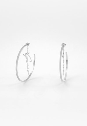 Silberne Creolen-Ohrringe mit einer zarten Kette, die ein kursives „Guess“-Logo in jedem Ring auf weißem Hintergrund aufhängt.