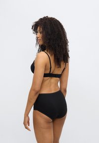 Μαύρο σουτιέν και ταιριαστό ψηλόμεσο σλιπ από λείο υλικό. Το σλιπ έχει κυματιστή άκρη. Το μοντέλο εμφανίζεται από πίσω.