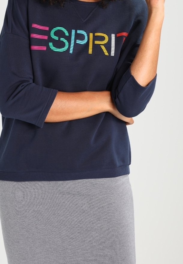 Mujer vestida con un sudadera azul marino con el texto colorido "ESPRIT" y una falda gris claro, con los brazos cruzados y una mano tocando su cara.
