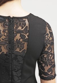 Svart klänning med en solid bakpanel och en spetsinsats, korta spetsärmar och dragkedjestängning bak. Strukturerade blommönster på spetsen.