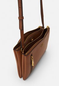 Ruskea teksturoitu synteettinen käsilaukku kultaisilla vetoketjuilla ja korostuksilla. Kaksi osastoa, säädettävä hihna ja logo edessä.