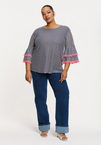Schwarz-weiß gemustertes Oberteil mit Glockenärmeln und rosa Pomponbesatz, kombiniert mit blauen weiten Jeans und silbernen Sandalen.