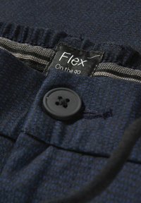 Tissu bleu marine avec un motif texturé, comprenant un bouton noir et une étiquette indiquant "Flex On the go." Détail d'une ceinture élastique grise.