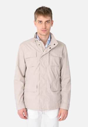 Giovane uomo che indossa una giacca beige con multiple tasche frontali e un colletto alto sopra una camicia blu, in piedi contro uno sfondo bianco.