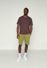 T-shirt marrone a maniche corte abbinato a pantaloni corti verdi, calze bianche e sneakers bianche. Design semplice con dettagli minimal di branding.
