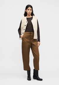 Ελεφαντόδοντο γιλέκο από μάλλινο ύφασμα με σκοτεινά καφέ κουμπιά πάνω από καφέ μπλούζα με μακριά μανίκια, συνδυασμένο με ψηλόμεσο καφέ παντελόνι και μαύρα μποτάκια στο ύψος του αστραγάλου.