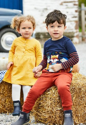 Twee kinderen zitten op een hooibaal. Het meisje draagt een gele jurk met stippen, en de jongen heeft een marineblauw-shirt met een vosontwerp en rode broek.