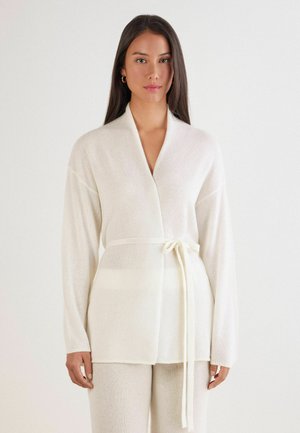 Cardigan en tricot blanc avec une taille ceinturée, des manches longues et une texture douce. Présente un col châle et des détails de bord subtils.