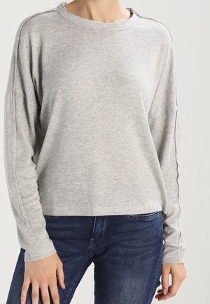 Felpa grigia chiaro, a maniche lunghe, con collo rotondo e spalle basse. Presenta una consistenza morbida e una vestibilità rilassata. Abbinata a jeans blu.