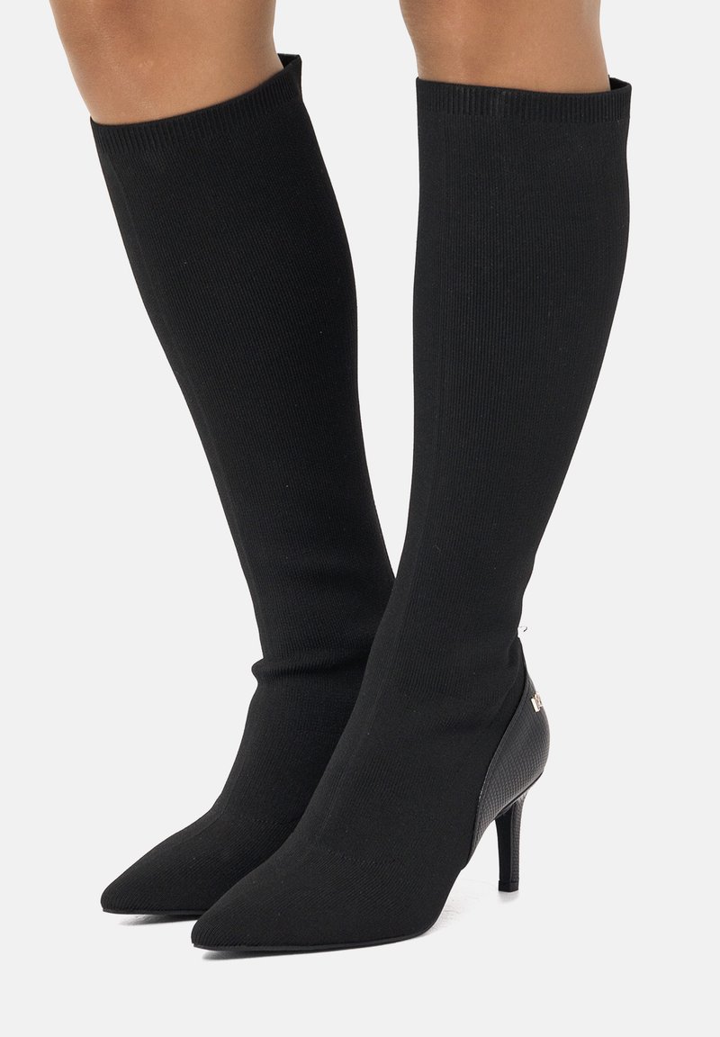 Botas de punto negras hasta la rodilla con punta afilada y tacón de aguja. Presentan un acento texturizado negro en el talón con un detalle de herrajes dorados.
