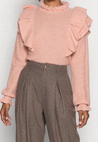 Světle růžový pletený svetr s volány na ramenou a manžetách, kombinovaný s hnědými kalhotami s vysokým pasem, které mají přední záhyby a zapínání na knoflík.