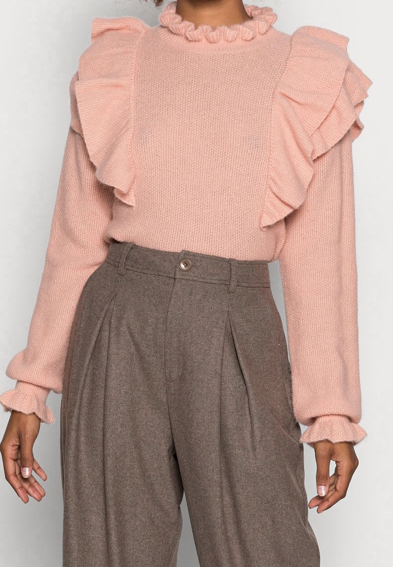 Světle růžový pletený svetr s volány na ramenou a manžetách, kombinovaný s hnědými kalhotami s vysokým pasem, které mají přední záhyby a zapínání na knoflík.