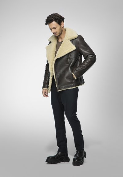 Giacca di pelle marrone con colletto in shearling, chiusura con zip e tasche laterali, abbinata a pantaloni neri e stivali neri robusti.