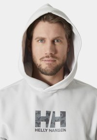 Sudadera blanca con un gran logo de camuflaje gris "HH" y "HELLY HANSEN" impreso debajo. Cuenta con una capucha con cordón y una textura suave.