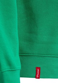 Felpa verde in cotone, con polsini e orlo a coste. Un'etichetta rossa con testo bianco è cucita in basso a sinistra.