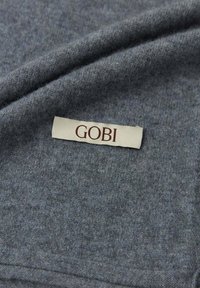 Grått stickat tyg med en mjuk textur, som visar en GOBI-etikett i krämfärg med mörkbrun text. Slät yta utan synliga mönster.