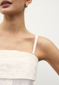 Haut en coton blanc avec bretelles ajustables, orné de broderies florales et de motifs texturés. Finition lisse, coupe semi-ajustée.