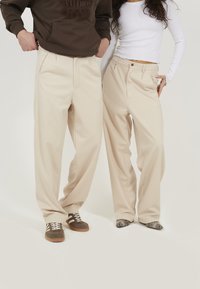 Pantalons beiges taille haute avec une coupe large, dot�és de plis à l'avant et de deux poches latérales. Texture lisse et design ajusté.