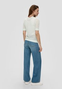 Biały sweter robiony na drutach z krótkim rękawem i teksturowanym wzorem, w połączeniu z jasnoniebieskimi spodniami o szerokich nogawkach i białymi butami na platformie.