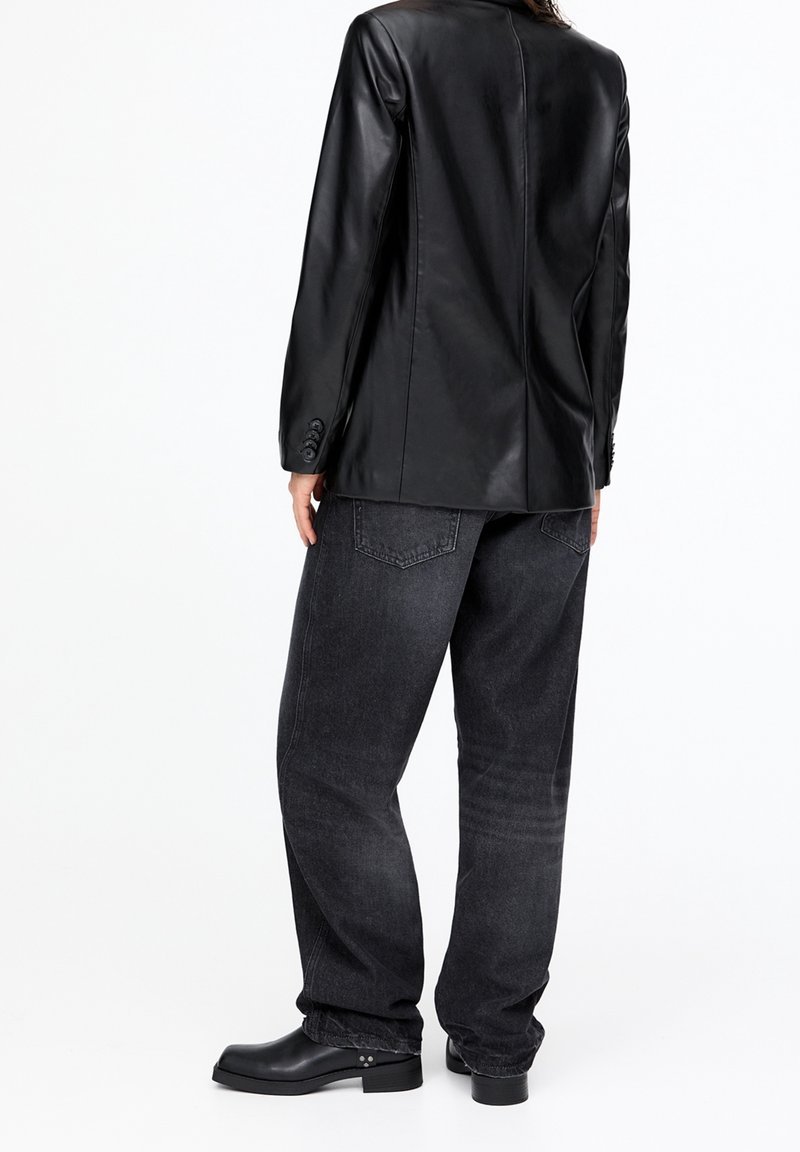 Blazer in pelle nera con una vestibilità sartoriale, chiusura a un bottone e spalle strutturate, abbinato a jeans a gamba larga grigio scuro e stivali neri.
