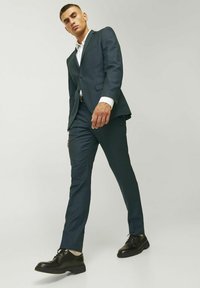 Tmavo modrý oblek s upraveným sako a úzkymi nohavicami. Biely dress shirt pod ním, doplnený o čierne šnurovacie topánky. Minimálne detaily.