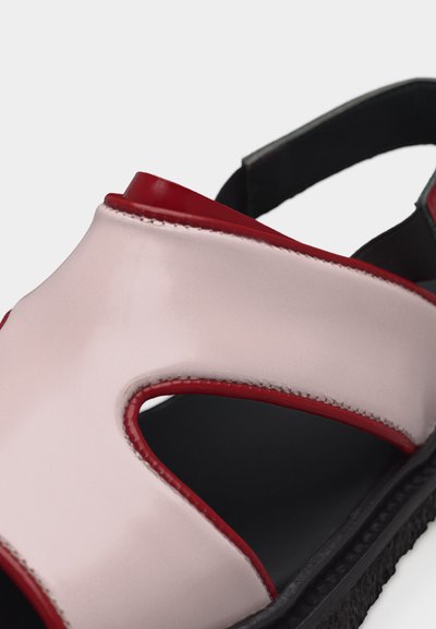 Κοντινό πλάνο ενός μαύρου σανδάλι με σόλα, με απαλές ροζ και κόκκινες δερμάτινες λουράκια και μια κομμένη λεπτομέρεια στο πάνω μέρος.