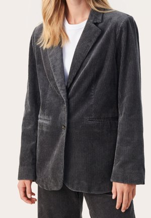 Femme portant un blazer en velours côtelé gris foncé et un pantalon assorti sur une chemise blanche, vue du cou jusqu'à mi-cuisse.