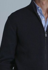 Marineblauer gerippter Cardigan mit durchgehendem Reißverschluss und hohem Kragen, aus einem glatten, strukturierten Stoff. Darunter befindet sich ein blau gestreiftes Hemd.