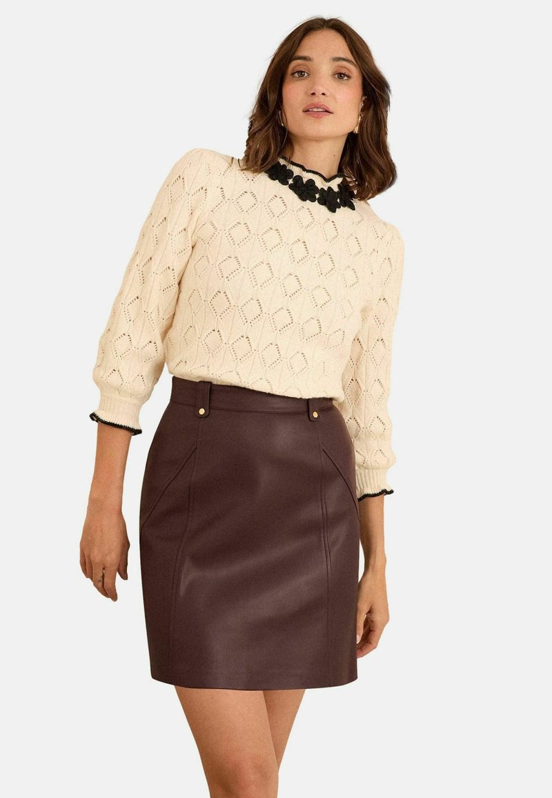 Krem pulover od pletene vune s dijamantnim uzorkom, crni cvjetni detalj na ovratniku. Burgundska suknja od umjetne kože s bočnim džepovima i zlatnim akcentima.