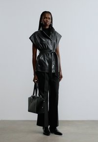 Zwart leren vest met brede kraag en een taille met ceintuur, gecombineerd met zwarte broek en een bijpassende handtas. Soepele textuur en een op maat gemaakte pasvorm.