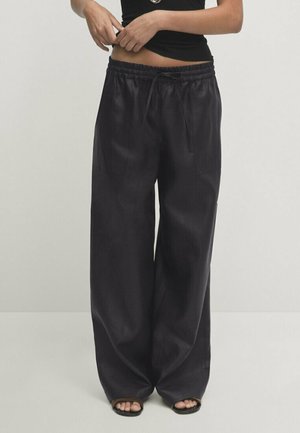 Osoba nosząca luźne, czarne, satynowe spodnie typu szerokie nogawki z elastycznym pasem i sznurkiem, w połączeniu z czarnymi sandałami na odkryte palce, regulująca czarną bluzkę.