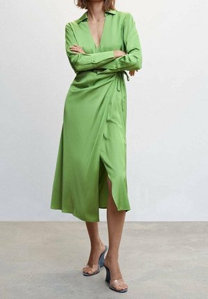 Femme les bras croisés portant une longue robe portefeuille verte et des sandales à talons avec brides transparentes, debout sur un sol gris contre un mur blanc.