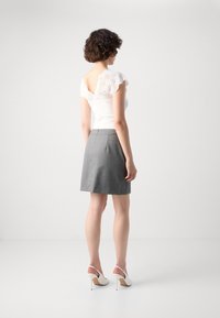 Haut blanc avec des accents en dentelle et une coupe ajustée, associé à une jupe grise. Le mannequin porte des chaussures à talons blancs, mettant en valeur une silhouette élancée.