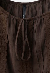 Vêtement en tissu marron avec panneaux en dentelle, encolure froncée et cordon noué au centre devant.
