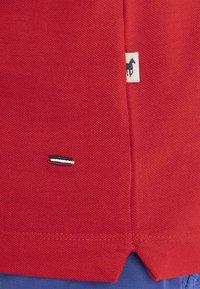 Hém de chemise en tissu texturé rouge avec un petit logo rayé bleu marine, blanc et rouge et une étiquette blanche arborant le profil d'un cheval noir.