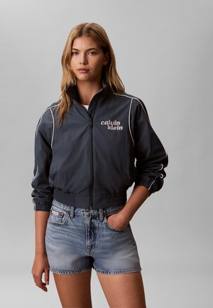 Ung kvinde iført en sort Calvin Klein jakke med lynlås og lyse denimshorts, stående med den ene hånd i lommen mod en grå baggrund.