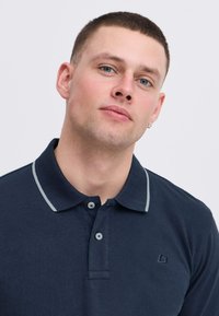 Mörkblå polo skjorta med krage, ljusgrå detaljer, två knappar och broderad logotyp på vänster bröst. Slät bomullstextur.