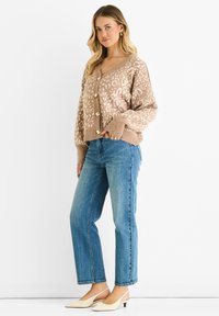 Beige knitted cardigan with abstract pattern, button closure, and ribbed cuffs, paired with blue straight-leg jeans and cream slingback shoes.