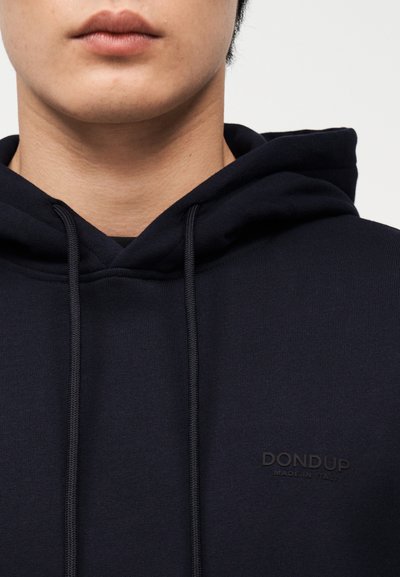 Κοντινό πλάνο ενός ατόμου που φοράει μαύρη μπλούζα με κουκούλα και κορδόνια, με διακριτικό κείμενο "DONDUP Made in Italy" στο στήθος.