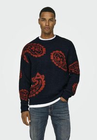 Marineblauwe trui met rode paisleypatronen, textuurstof, ronde hals en geribde manchetten; gedragen over een witte shirt, gecombineerd met blauwe jeans.