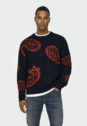 Marineblauwe trui met rode paisleypatronen, textuurstof, ronde hals en geribde manchetten; gedragen over een witte shirt, gecombineerd met blauwe jeans.