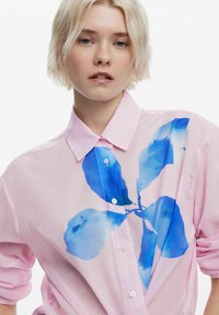 Camicia rosa con bottoni caratterizzata da una stampa floreale blu ad acquerello sul davanti. Realizzata in un tessuto morbido con colletto classico e maniche ampie.
