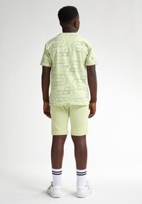 Lichtgroen shirt met korte mouwen en bijpassende shorts met zwarte geometrische patronen. Witte sokken met zwarte strepen en witte sneakers.