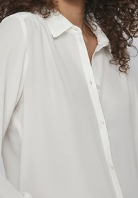 White, lightweight blouse with a button-down front, classic collar, and soft texture. Features long sleeves and pleated detailing at the shoulders.