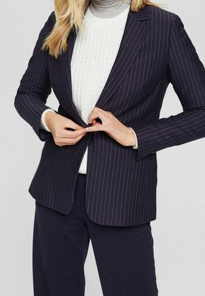 Person, der knappede en mørk stribet blazer over en hvid struktureret sweater på, iført mørke bukser mod en ensfarvet lys baggrund.