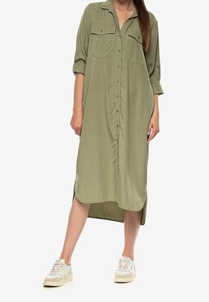 Vrouw die een olijfgroene overhemdjurk met opgerolde mouwen draagt, gecombineerd met beige-witte sneakers, staand met één uitgestrekt been.