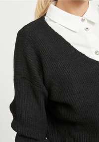 Czarny sweter z dzianiny, z dekoltem w kształcie litery V, grubą teksturą ribbed oraz luźnym krojem, założony na białą koszulę z kołnierzykiem z guzikami.