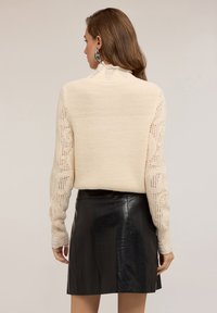 Světle krémový pletený svetr s ribkovanou strukturou a otevřenými pletenými rukávy, doplněný černou koženou sukní. Na svetr jsou viditelné roztrhané detaily.