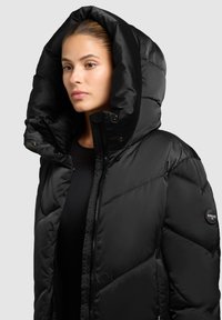 Schwarze Steppjacke mit großer Kapuze, mit einem gesteppte Design und einem runden Logo am Ärmel. Glatte, glänzende Textur.