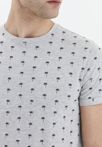 Grå kortærmet t-shirt med rund halsudskæring, prydet med et gentaget sort palmetræsmønster, lavet af blødt, tekstureret stof.